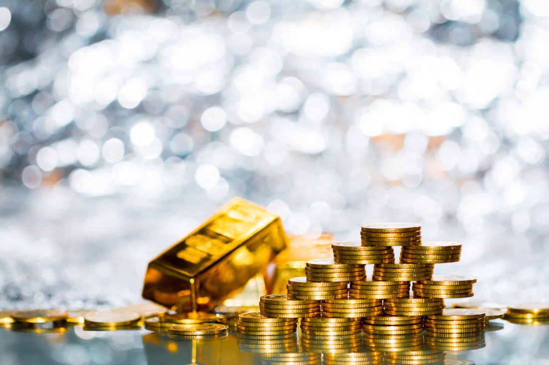 Stacked gold coins arranged in pyramid formation beside a shiny gold bullion bar on reflective surface