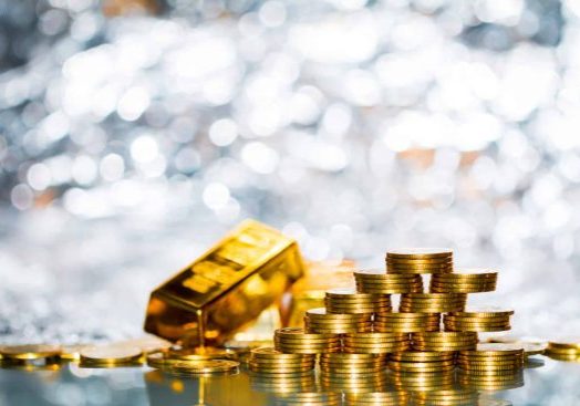 Stacked gold coins arranged in pyramid formation beside a shiny gold bullion bar on reflective surface